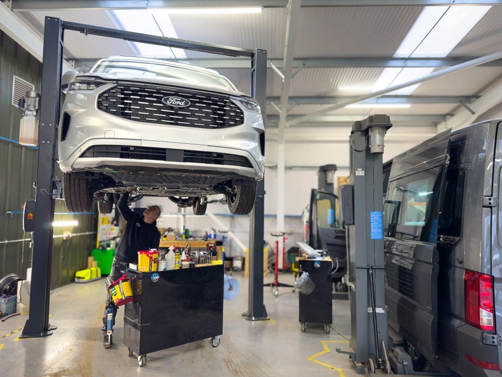 A service engineer underneath a white Ford Transit Custom campervan on ramps.