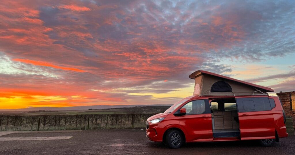 A red campervan with a pop top roof elevated with a beautiful sunrise in in front of farmland.
